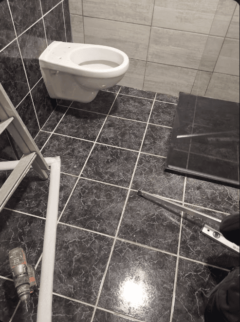 Toilette suspendue blanche sur mur gris, sol en carrelage noir marbré et outils de chantier.