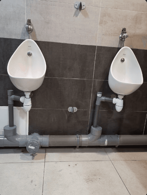Deux urinoirs blancs fixés au mur avec une tuyauterie de drainage grise apparente au sol.