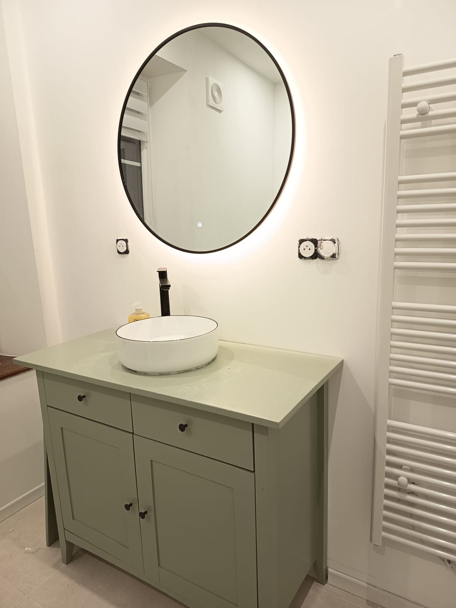 Salle de bain moderne avec meuble vert, vasque blanche et miroir rond rétroéclairé.