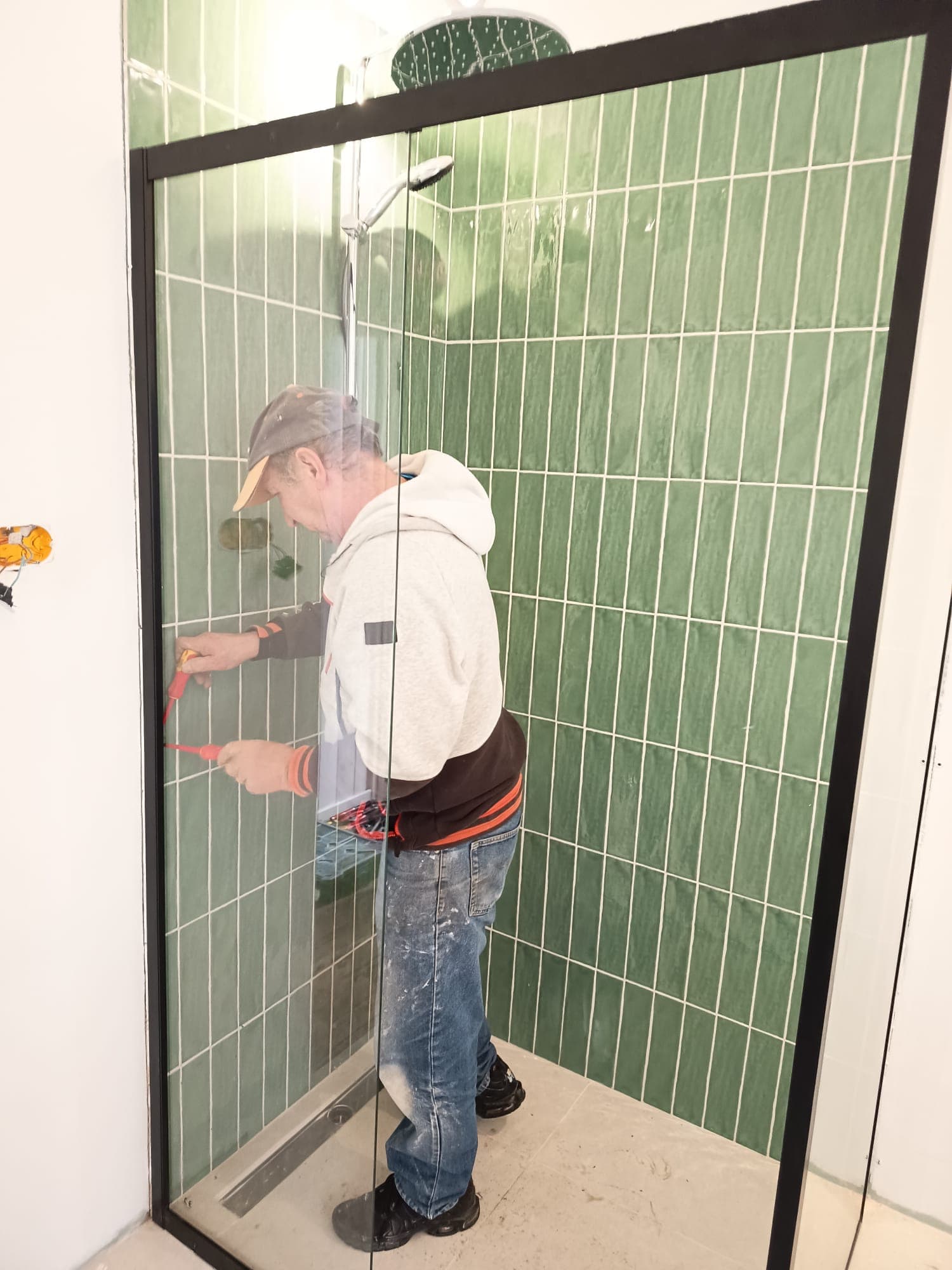 Un homme installe une porte de douche vitrée à cadre noir sur du carrelage vert.