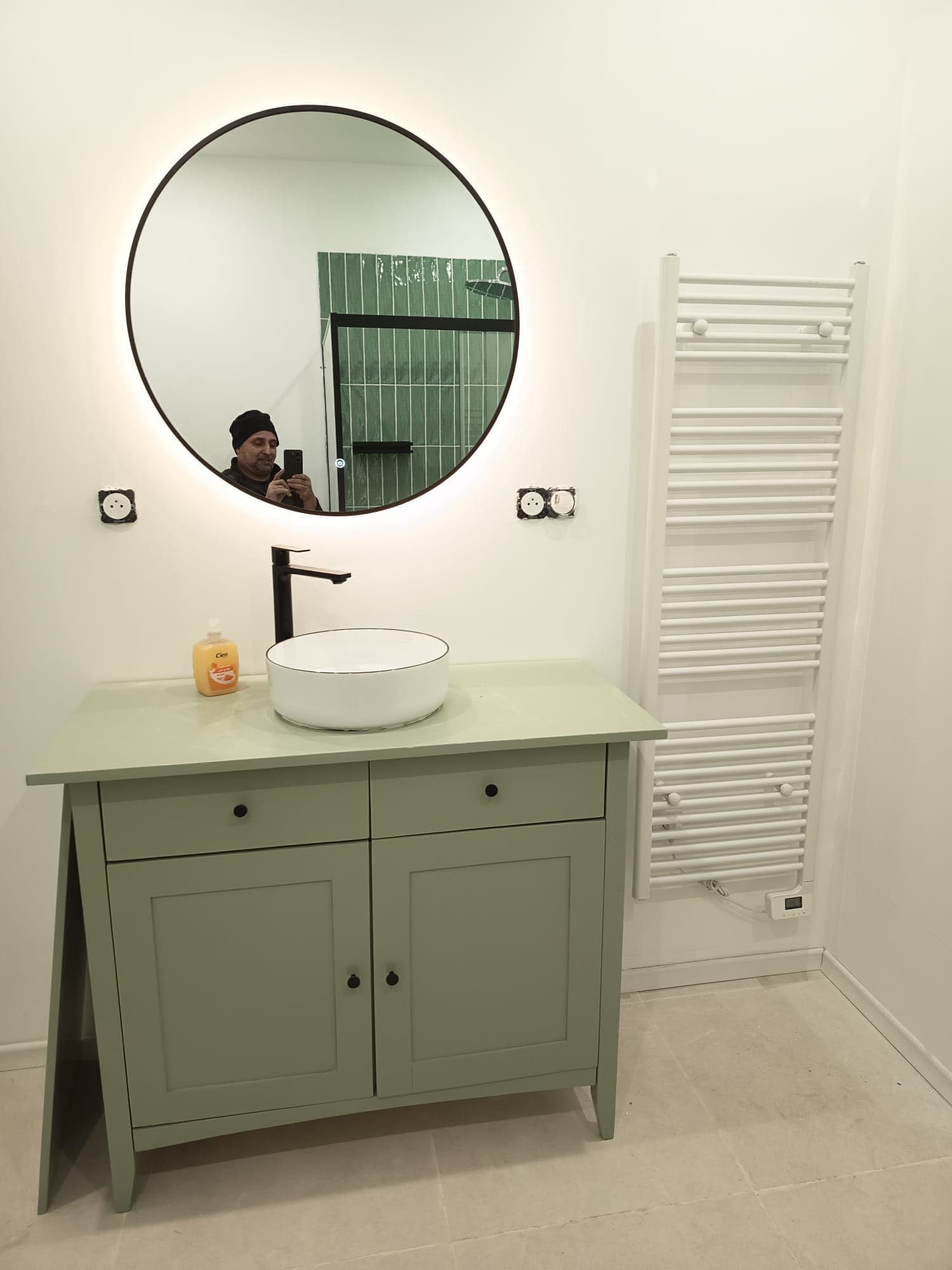 Meuble vasque vert sauge, robinet noir mat, miroir rond rétroéclairé et sèche-serviettes blanc.